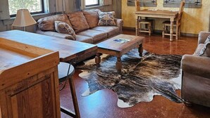 A rustic interior with a wooden ceiling, a ceiling fan, a leather sofa, a dining table with chairs, and a large window.