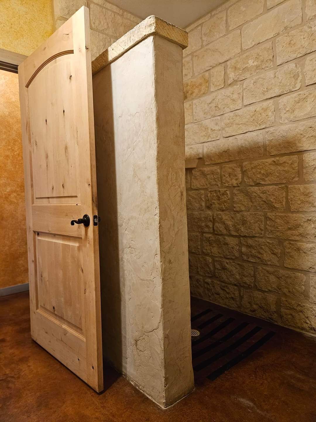 Una puerta de madera con un picaporte negro, entreabierta, muestra una pared de piedra con un bajante.
