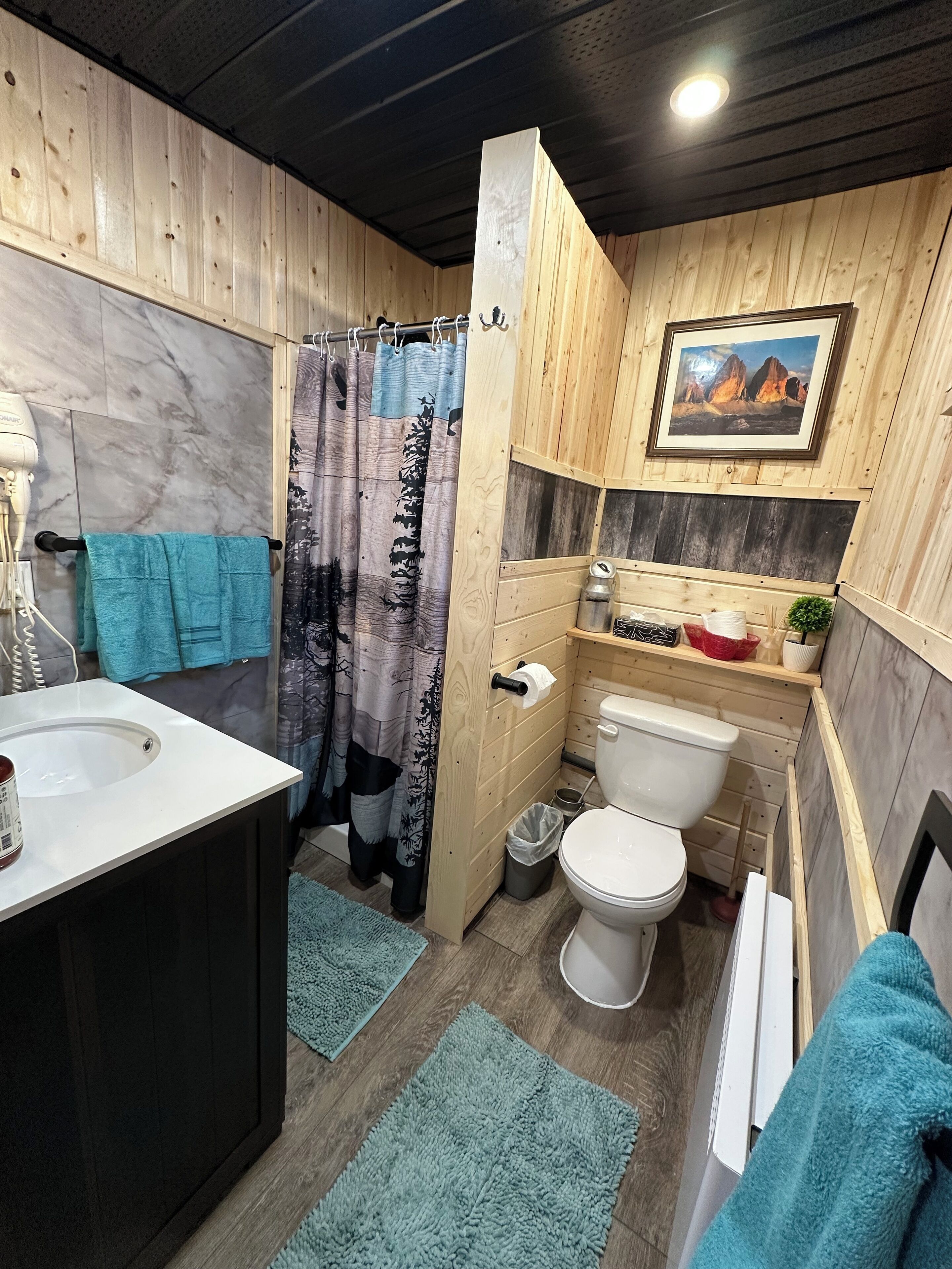 Un baño pequeño con revestimiento de madera, una pared de mármol, cortina de ducha con diseño de bosque, inodoro, lavabo y un estante pequeño con artículos de aseo.