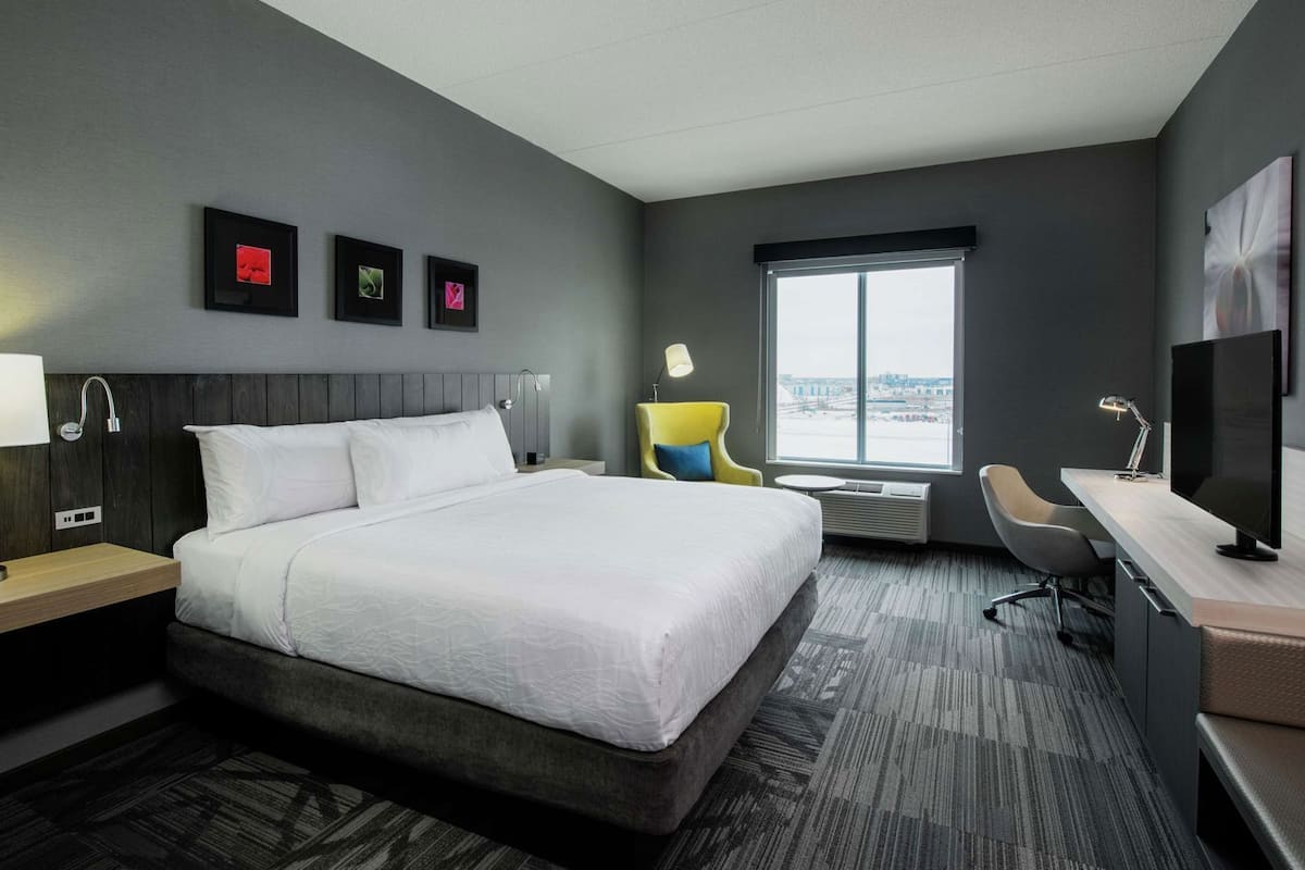 A hotel room with a bed, a desk, a chair, a desk lamp, a TV, a window, and artwork on the wall.
