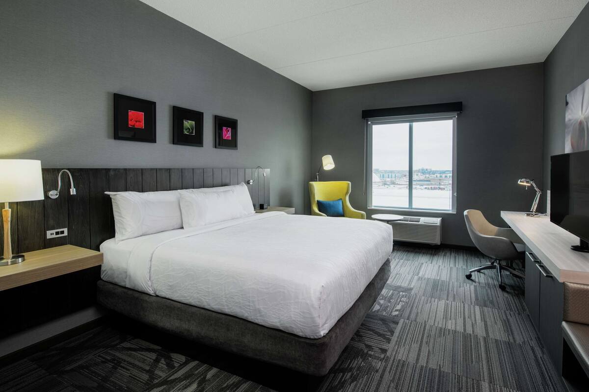 A modern hotel room with a large bed, a desk, a chair, and a window with a city view.