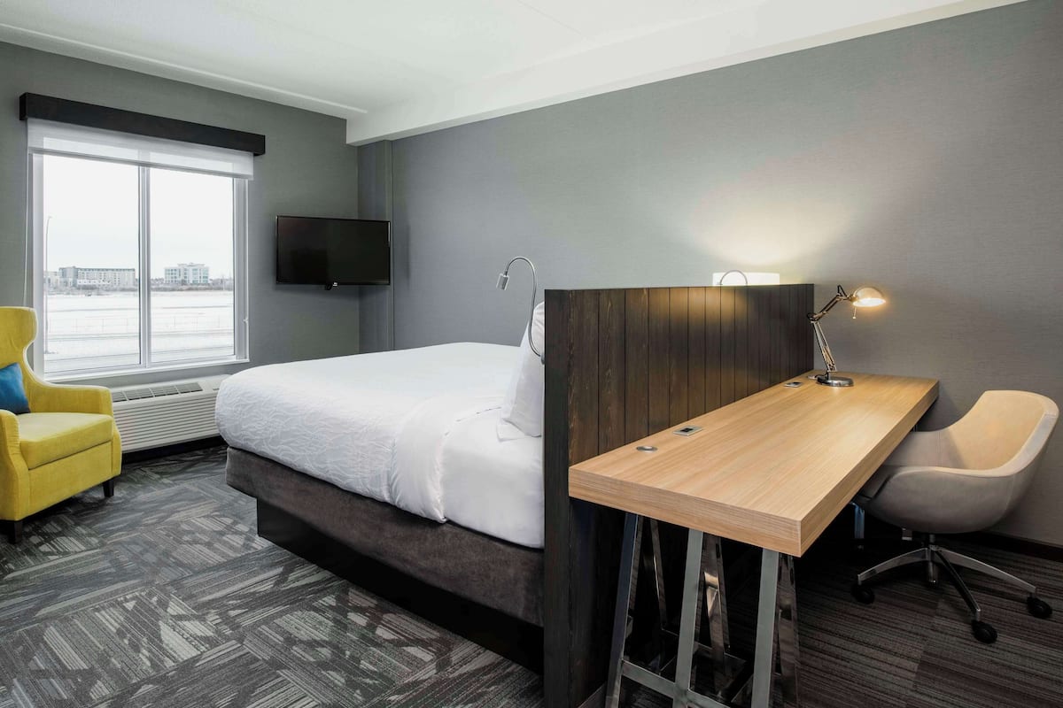A hotel room with a bed, a desk, a chair, a TV, and a window with a view of buildings.