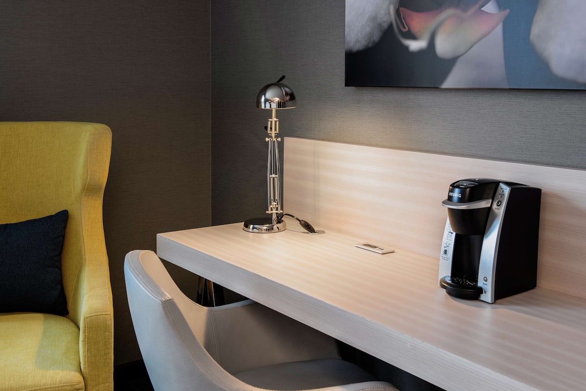 A hotel room with a desk, a modern lamp, a coffee maker, a yellow armchair, and a painting on the wall.