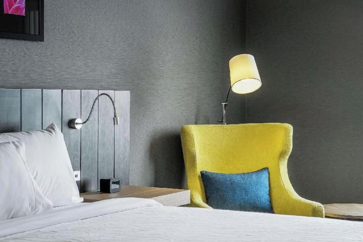 Une chambre d’hôtel avec un lit, une table de chevet, une lampe et un fauteuil jaune avec un coussin bleu.