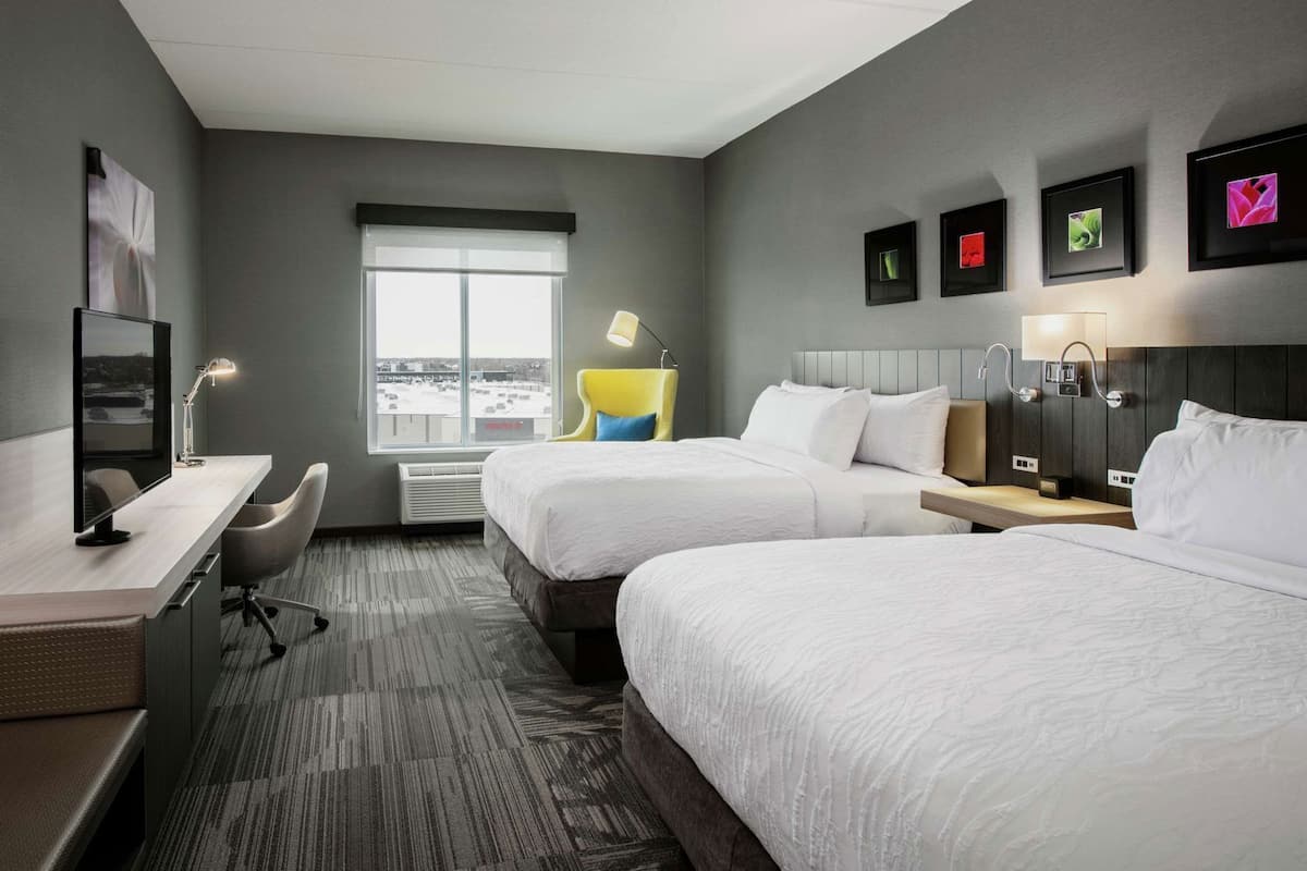 A hotel room with two beds, a desk, a chair, a TV, and a window with a view.