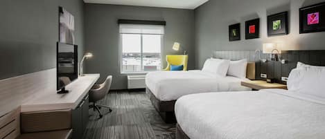 A hotel room with two beds, a desk, a chair, a window with a view, and framed pictures on the wall.