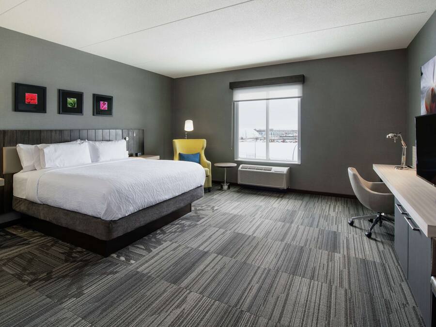 A hotel room with a large bed, a desk, a chair, a TV, and a window with a city view.