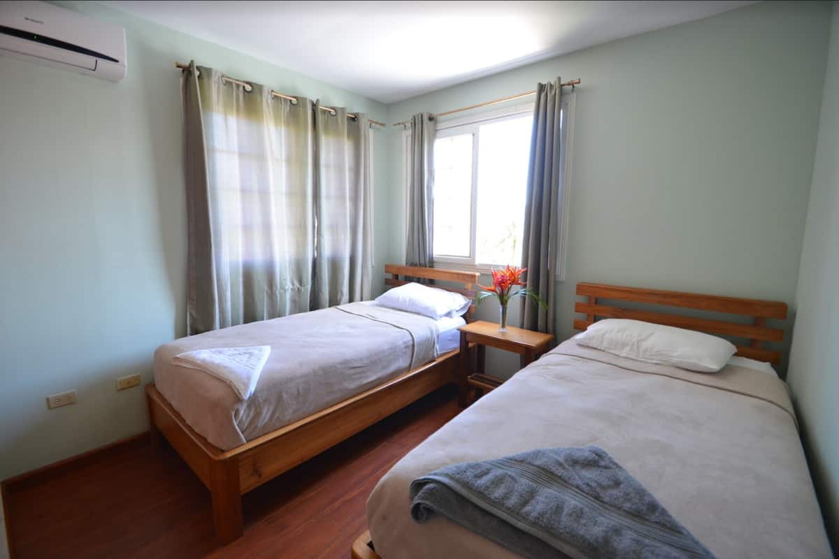 A room with two single beds, a wooden headboard, a nightstand with a vase of flowers, and a window with curtains.