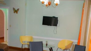 A round glass table with four chairs, a flat-screen TV mounted on the wall, and a chandelier. - Caribbean Villages Apartments (Bocas del Toro)
