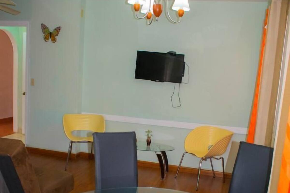 A round glass table with four chairs, a flat-screen TV mounted on the wall, and a chandelier.
