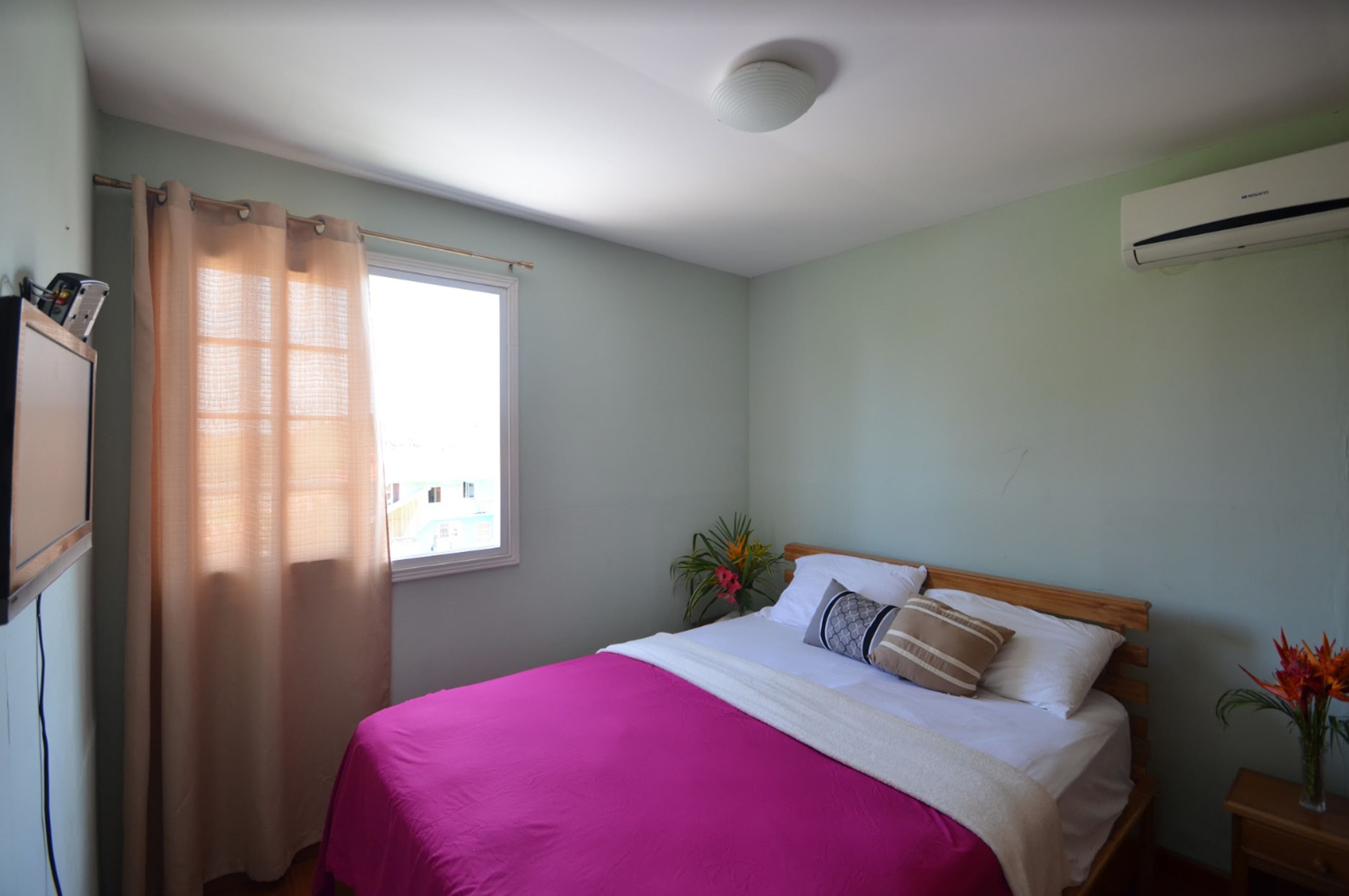 A bedroom with a bed, a window with a curtain, an air conditioner, and a nightstand with a vase of flowers.