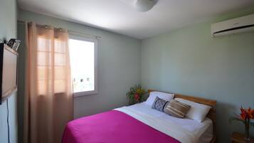 A bedroom with a bed, a window with a curtain, an air conditioner, and a nightstand with a vase of flowers.