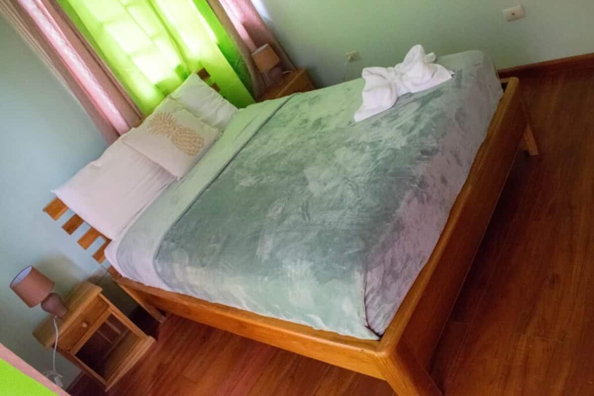 A single bed with a wooden frame, a green patterned bedspread, and white pillows. There is a small wooden nightstand with a lamp beside the bed.