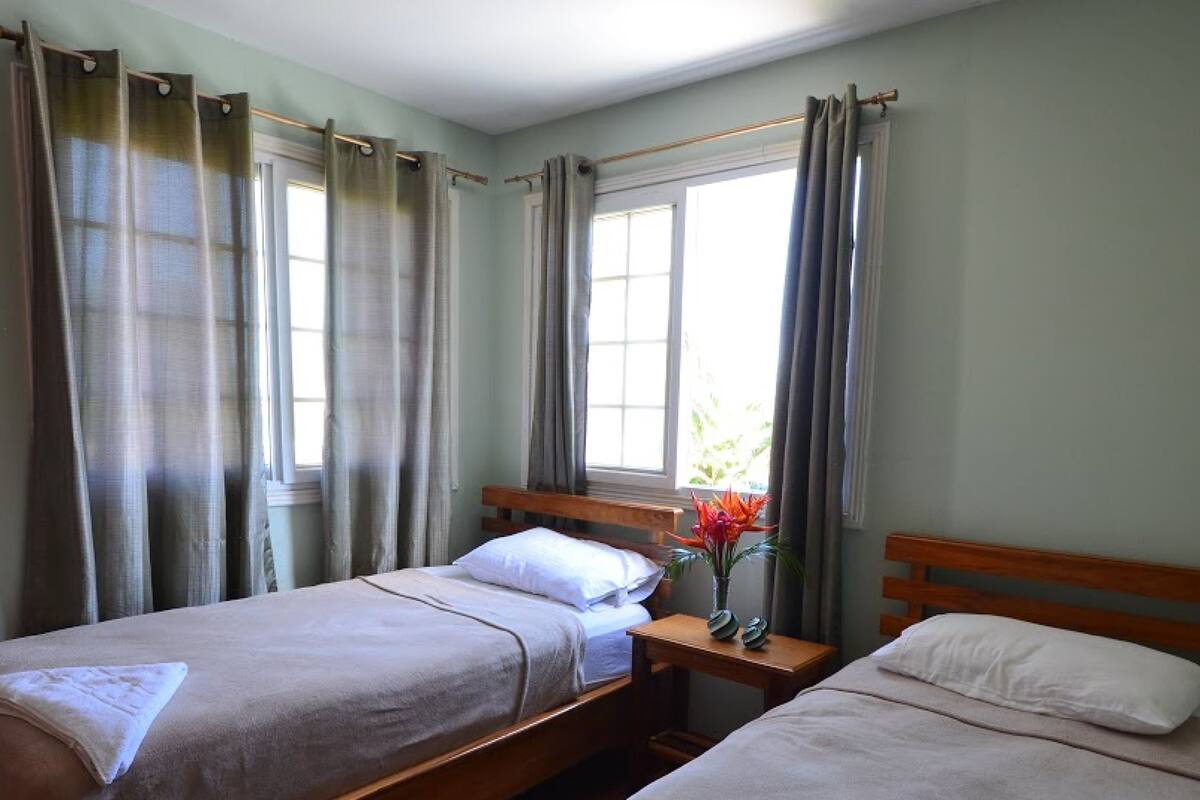 A room with two single beds, a window with curtains, and a small bedside table with a vase of flowers.