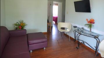 Una sala de estar con un sofá morado, una mesa de centro de vidrio, una silla blanca y un televisor montado en la pared.