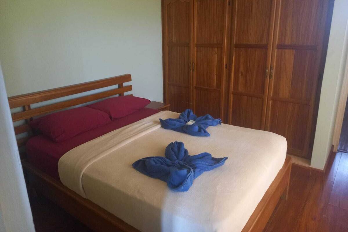 A bedroom with a wooden bed frame, a red pillow, and a beige bedspread. There are two blue towels on the bed.