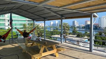 Una terraza al aire libre con una mesa y sillas de picnic de madera, una hamaca y vistas de un horizonte urbano sobre una masa de agua.