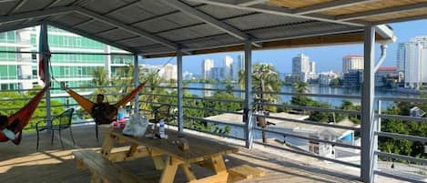 An outdoor deck with a wooden picnic table and chairs, a hammock, and a view of a city skyline across a body of water.