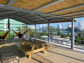 An outdoor deck with a wooden picnic table and chairs, a hammock, and a view of a city skyline across a body of water. - Casa Santurce - Hostel (San Juan)