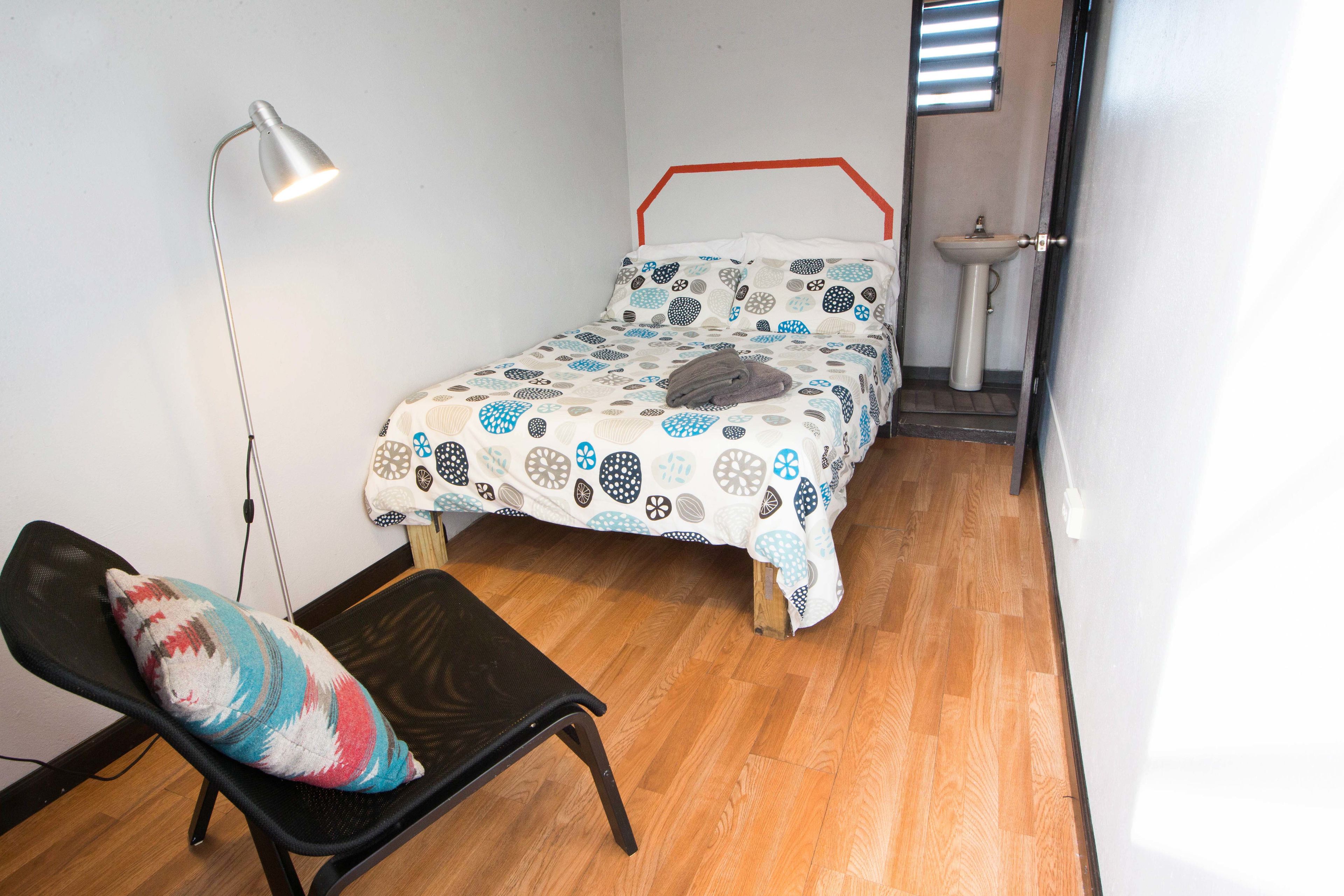 Ein kleines Zimmer mit einem Einzelbett, Holzboden, Nachttisch, Stuhl, Schreibtischlampe und einem Fenster mit Jalousien.