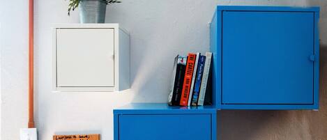Una habitación con un armario blanco, un armario azul y una planta pequeña encima. Hay libros en el armario azul y un cartel en la pared.