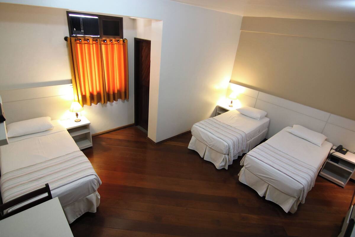A hotel room with three single beds, wooden flooring, and a window with orange curtains.