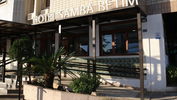 Un hotel llamado "HOTEL SAMBA BETIM" con fachada de baldosas, amplias ventanas y un área de entrada cubierta con palmeras.