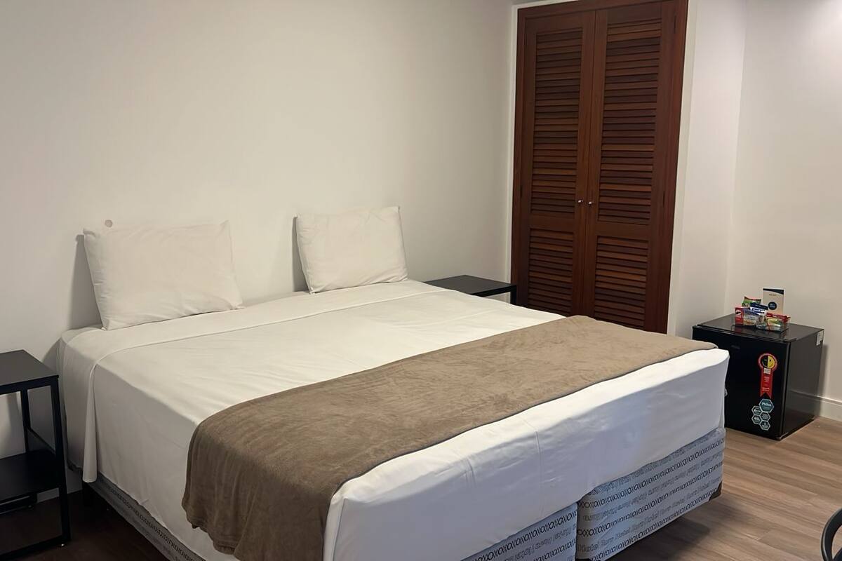 A neatly made bed with a white and brown blanket, a small bedside table, a wardrobe, and a compact refrigerator on the floor.
