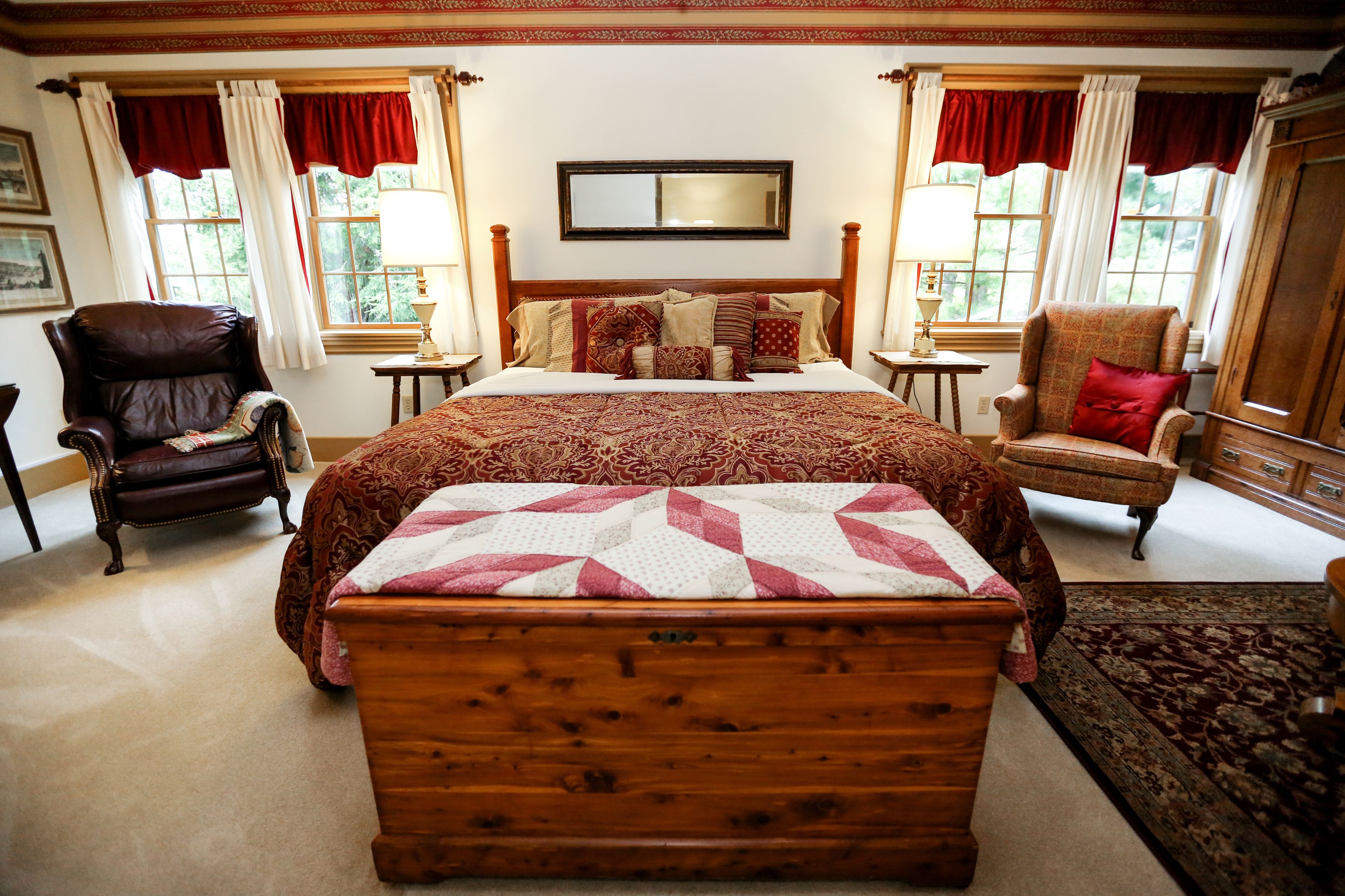 Um quarto espaçoso com uma cama de madeira grande, duas poltronas, um aparador de madeira e uma janela ampla com cortinas.