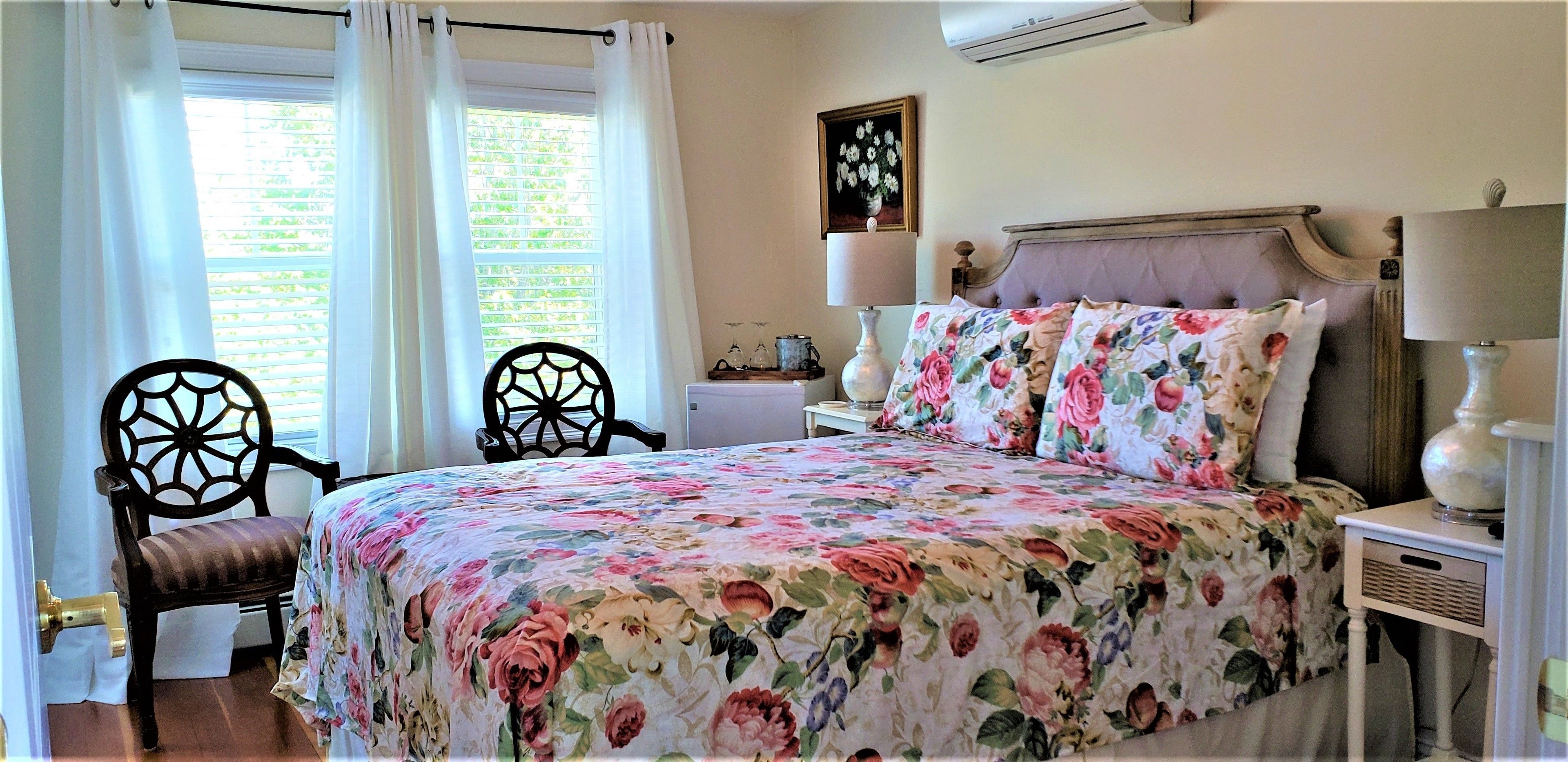 A bedroom with a floral bedspread, two chairs, a bedside table with a lamp, and a window with curtains.