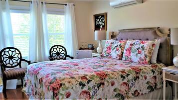 A bedroom with a floral bedspread, two chairs, a bedside table with a lamp, and a window with curtains.