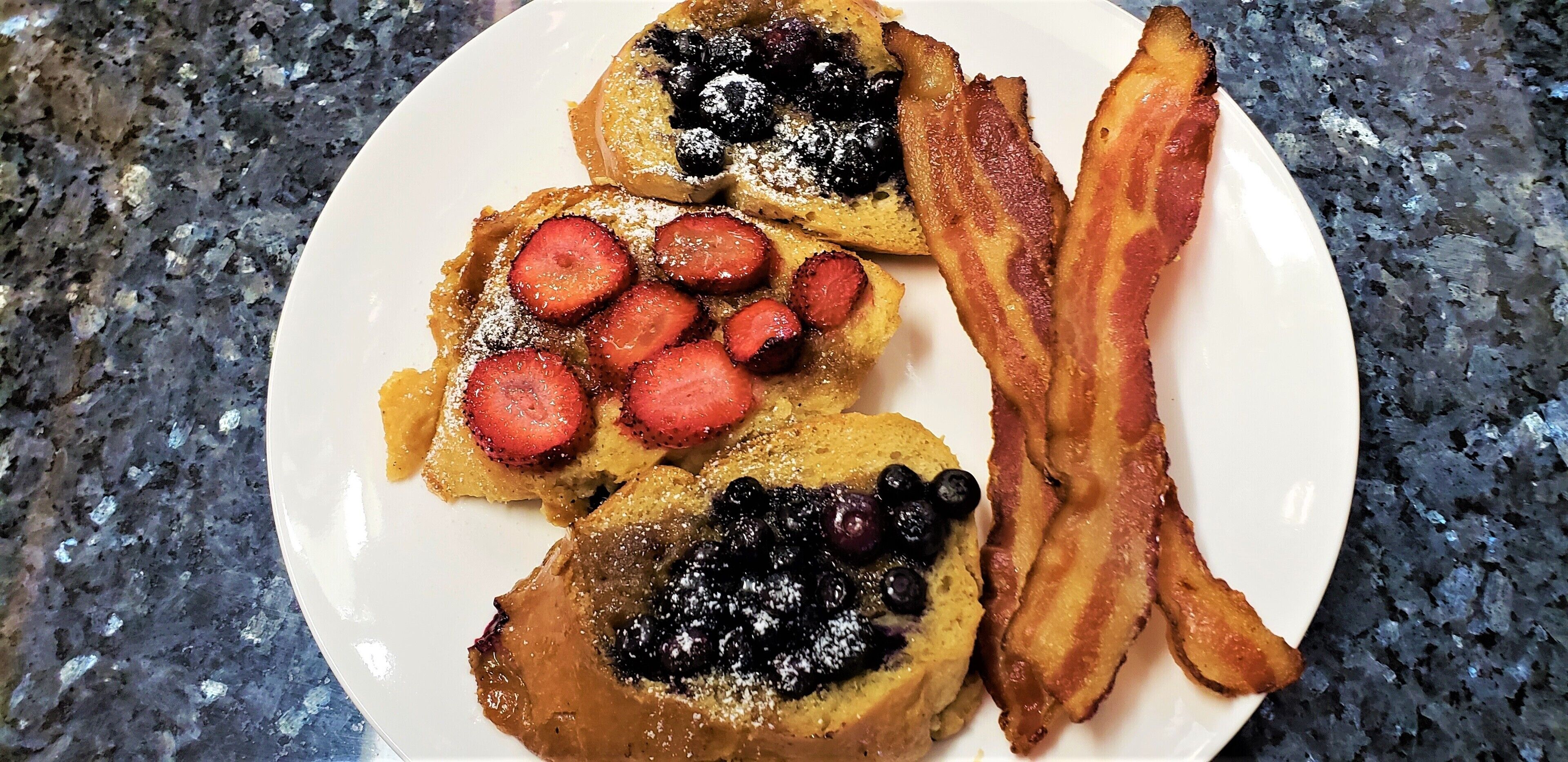 A plate with French toast topped with blueberries and strawberries, accompanied by a strip of bacon.