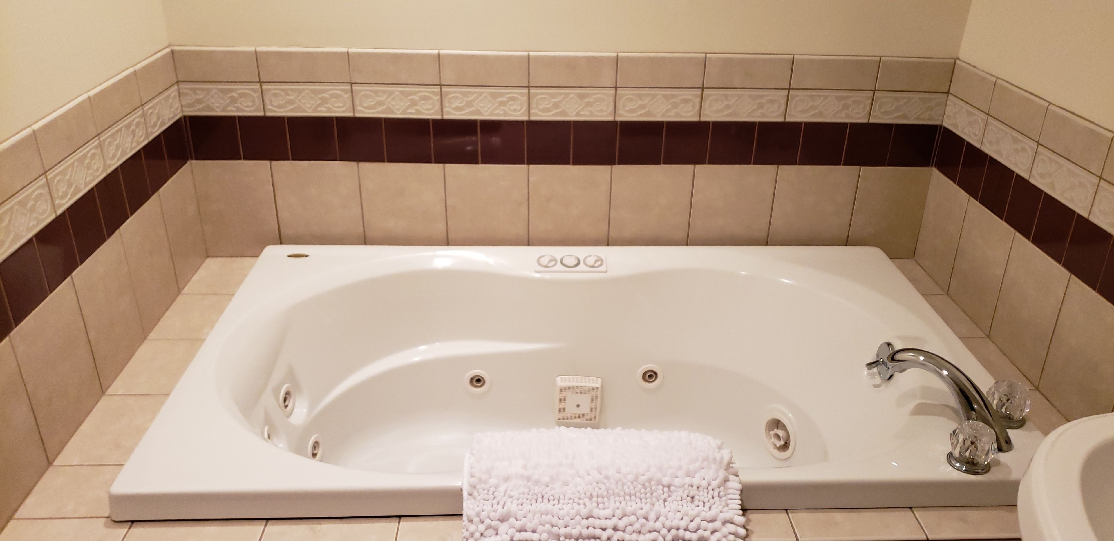 A large, rectangular bathtub with a single faucet and a small control panel, surrounded by beige and dark brown tiles.