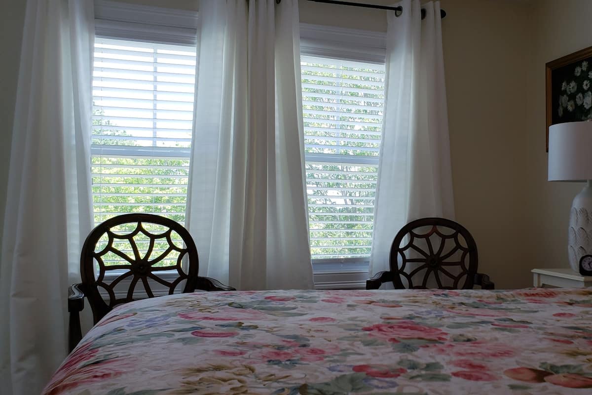 Un dormitorio con edredón estampado con motivos florales, cabecero de madera, mesita de noche con lámpara y ventana con persianas.
