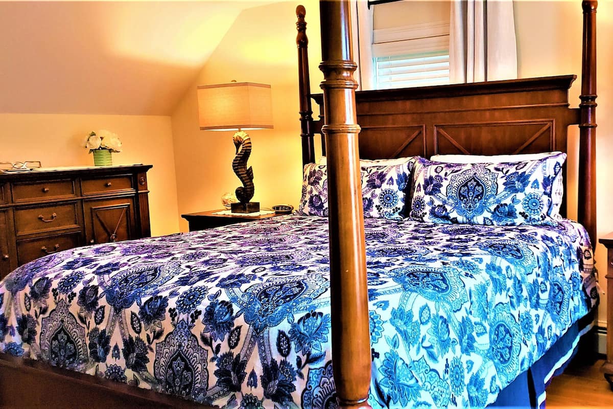 Una cama con dosel adornada con una colcha blanca y azul estampada, una mesita de noche de madera con una lámpara y un aparador con cajones.