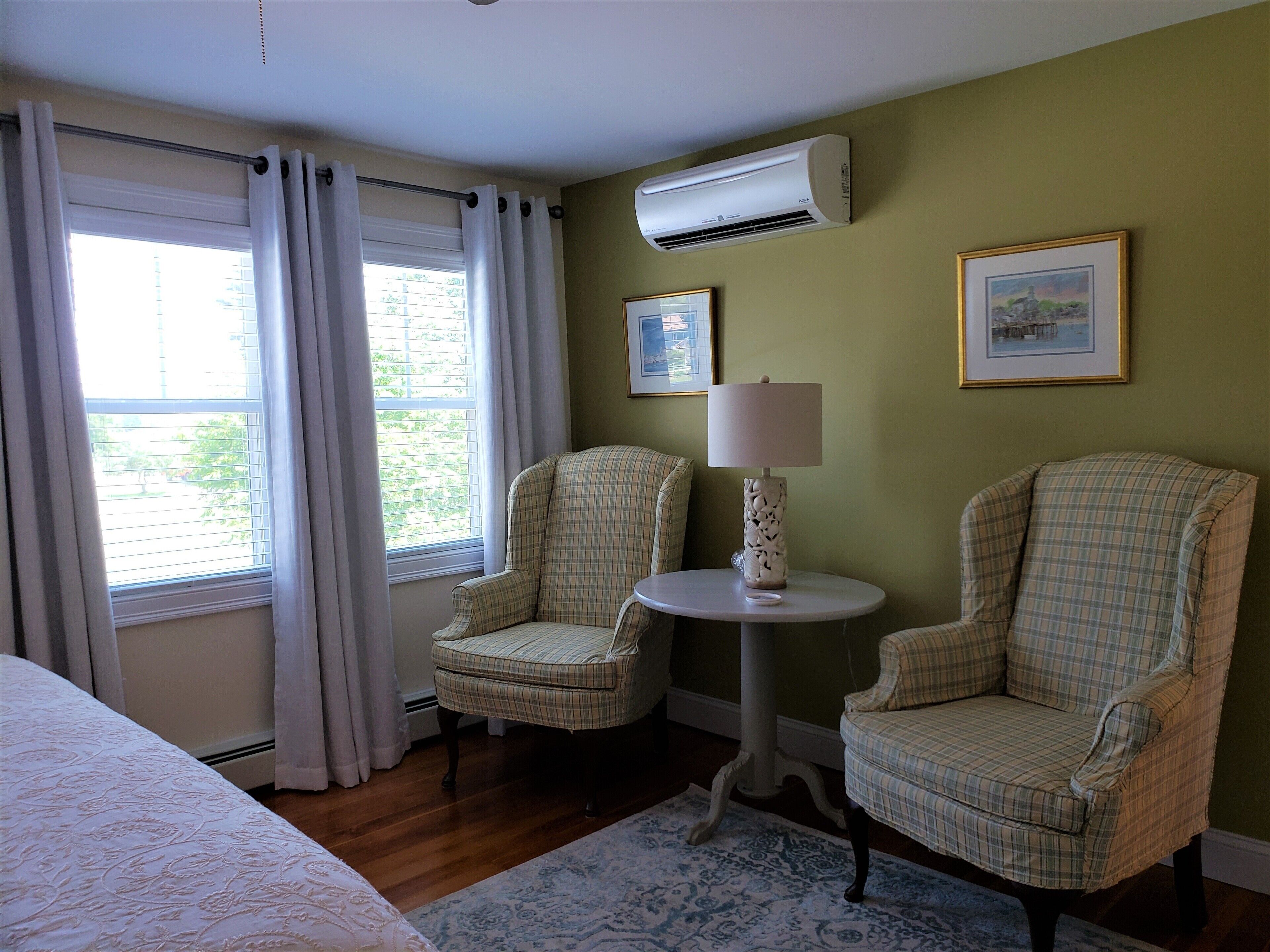 A hotel room with two plaid chairs, a round table with a lamp, an air conditioner, and a framed picture on the wall.