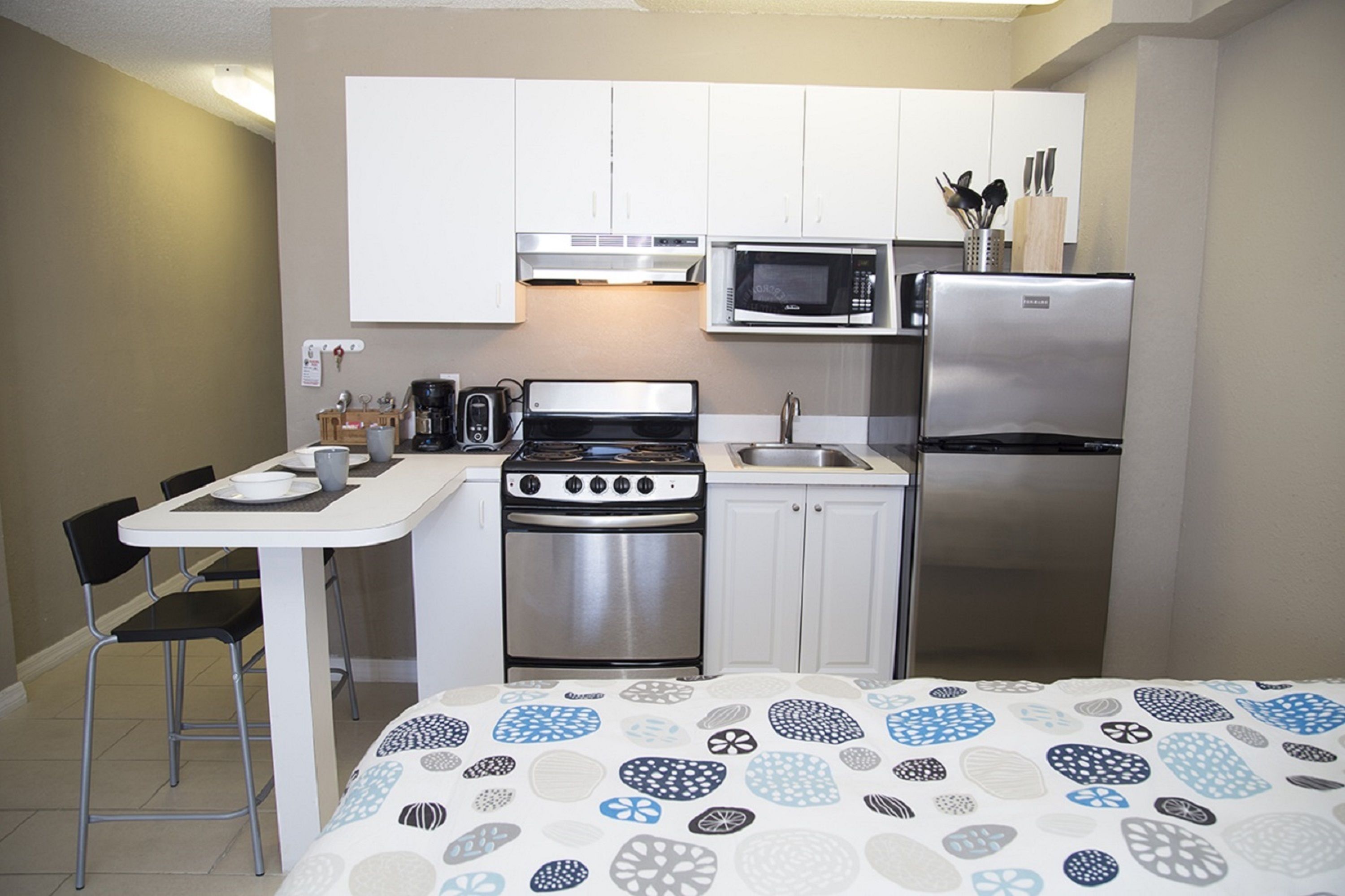 A compact kitchen with white cabinets, a stainless steel stove, a microwave, and a refrigerator. There is a small dining table with chairs and a bed with a patterned bedspread.