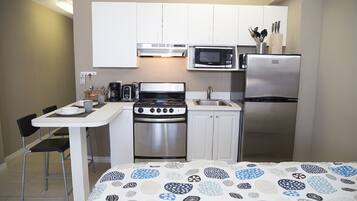 A compact kitchen with white cabinets, a stainless steel stove, a microwave, and a refrigerator. There is a small dining table with chairs and a bed with a patterned bedspread.