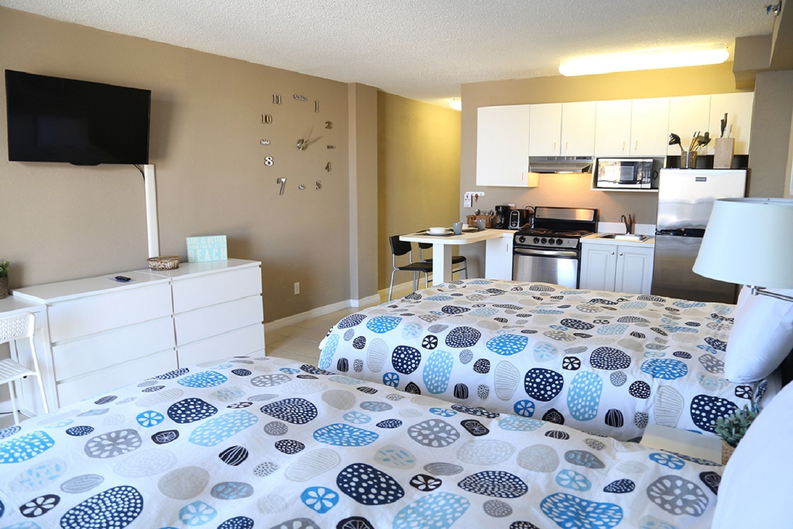 Habitación de hotel compacta con kitchenette, televisor de pantalla plana en la pared y cama con ropa de cama estampada.