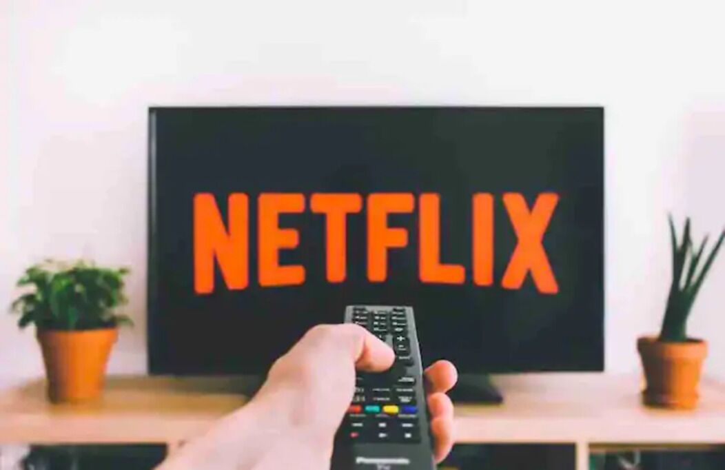 A hand holding a remote control in front of a TV with the Netflix logo.