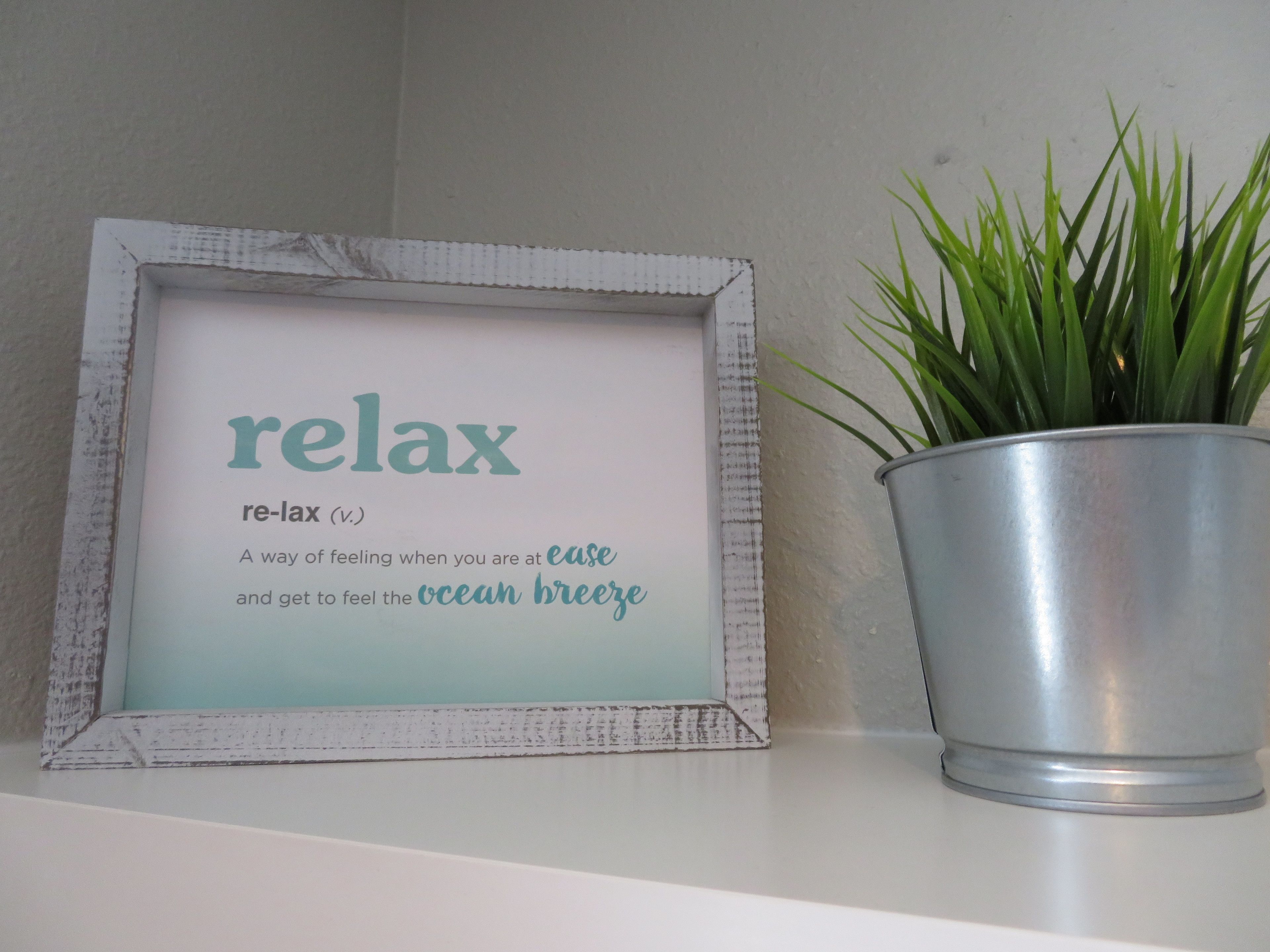 Une pancarte encadrée portant le mot «relax» et sa définition, placée à côté d’une plante en pot sur une étagère.