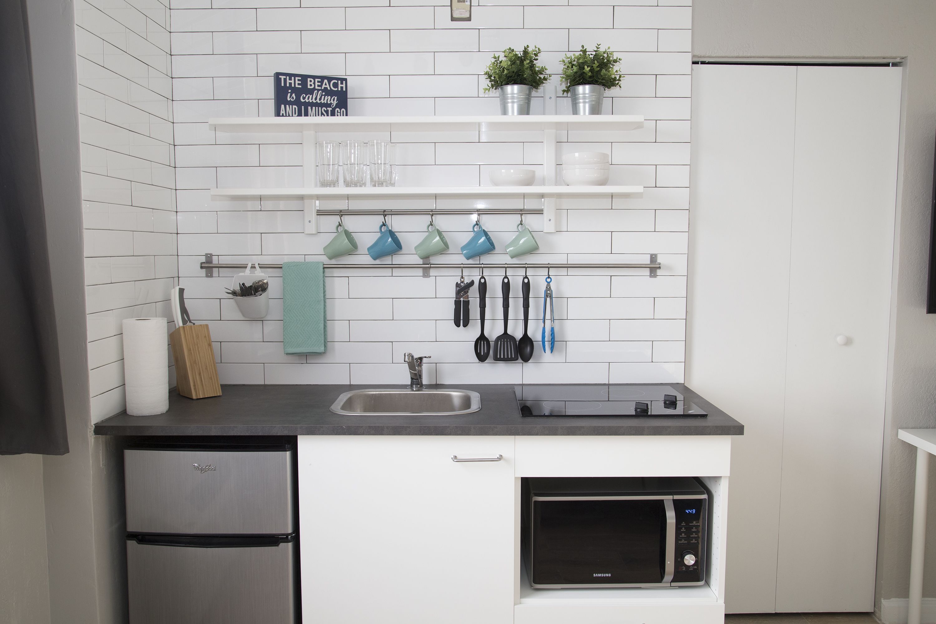A modern kitchen with white tiled backsplash, stainless steel appliances, and a shelf with kitchen utensils and plants.