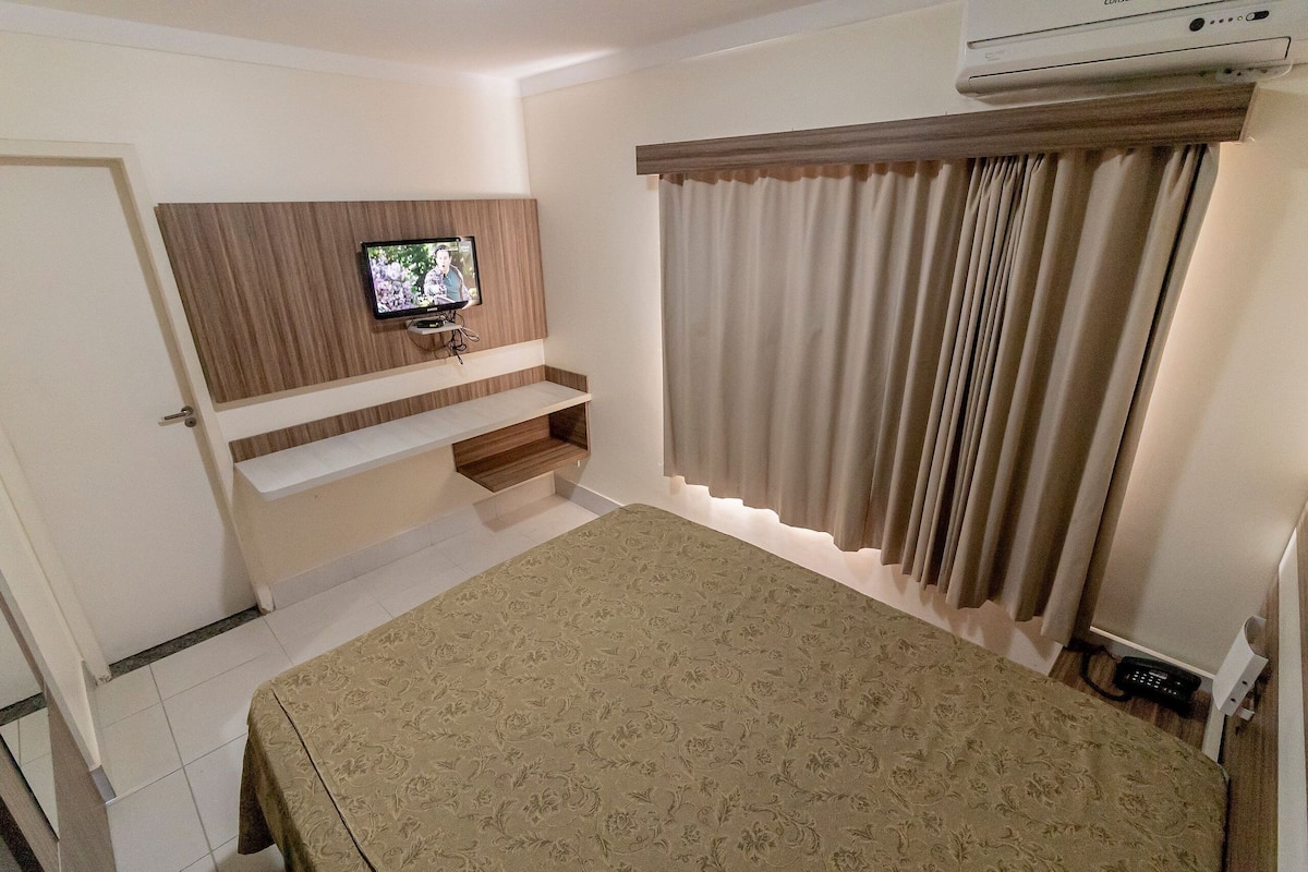 Habitación de hotel con cama, televisor montado, estante empotrado y ventana con cortinas.