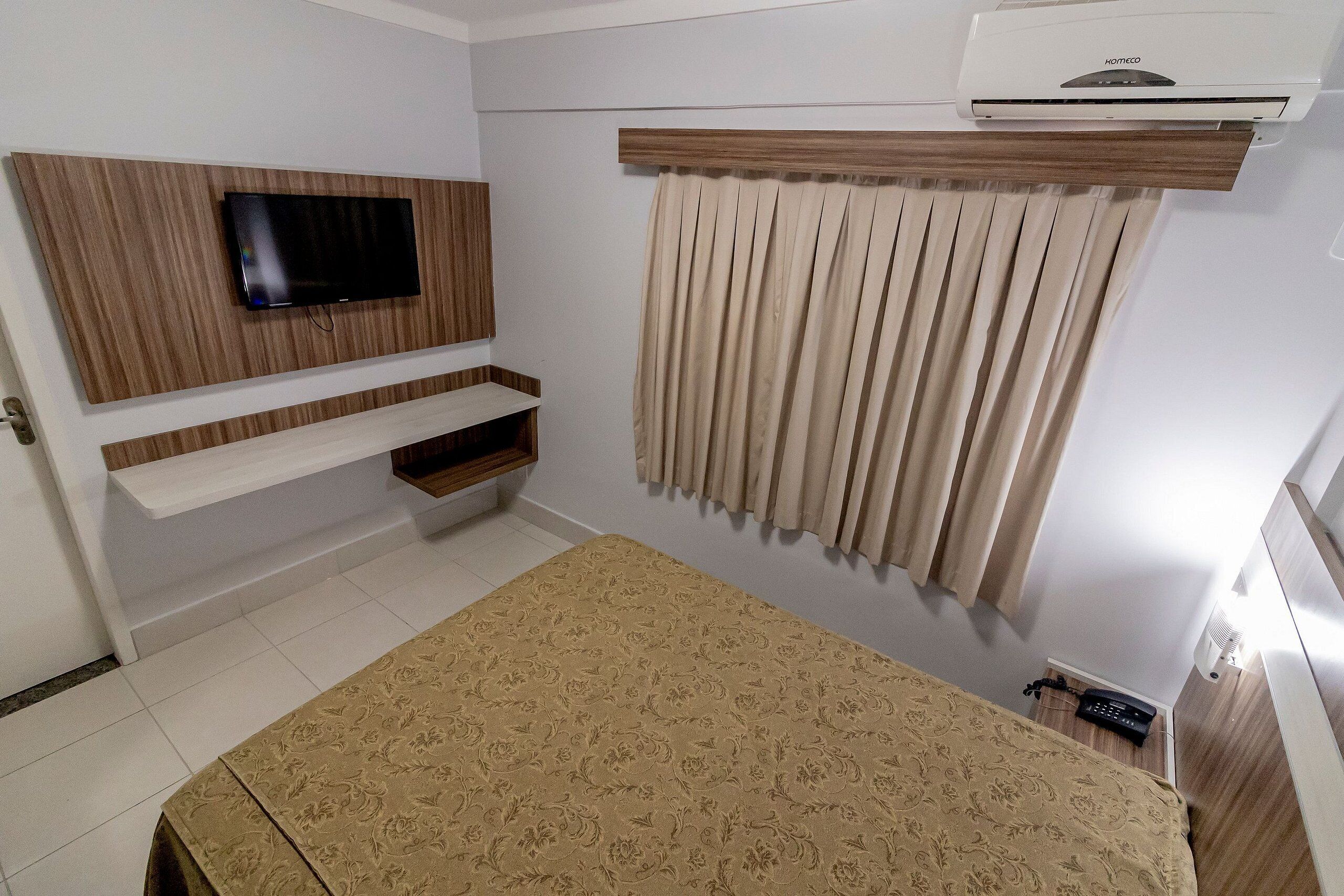Habitación de hotel con cama, televisor montado, estante empotrado, ventana con cortinas y aire acondicionado.