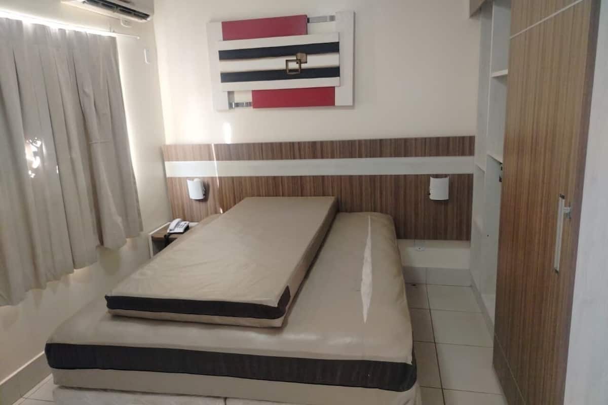 Una habitación de hotel con una cama, aire acondicionado instalado en la pared, una cortina, una mesita de noche con un teléfono y un armario de madera.