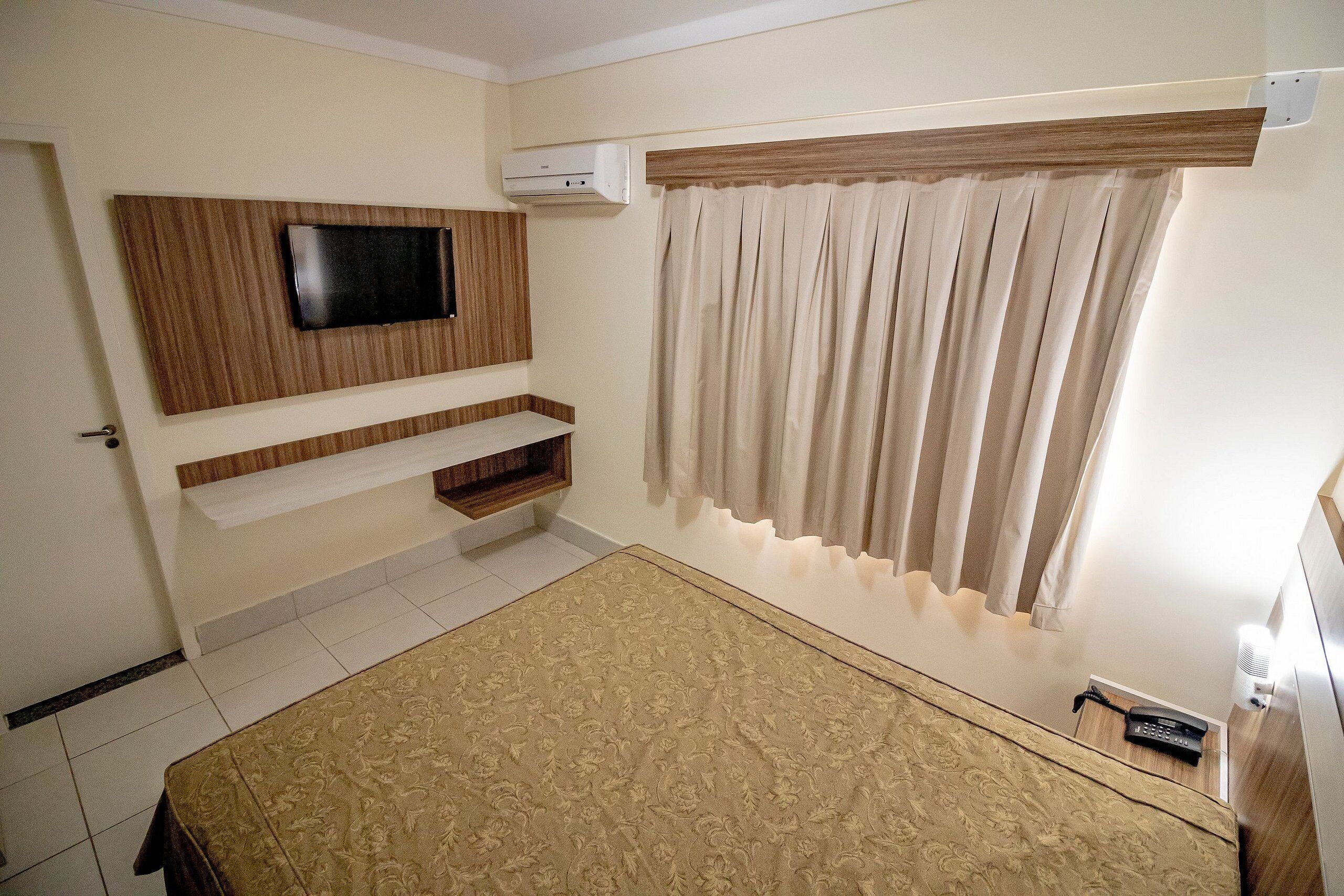 Quarto com cama, televisão montada na parede, prateleira embutida, janela com cortinas e aparelho de ar condicionado.