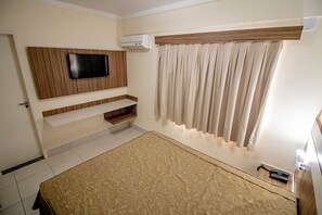 Une chambre à coucher comprenant un lit, un téléviseur fixé au mur, une étagère intégrée, une fenêtre avec des rideaux et un appareil de climatisation.