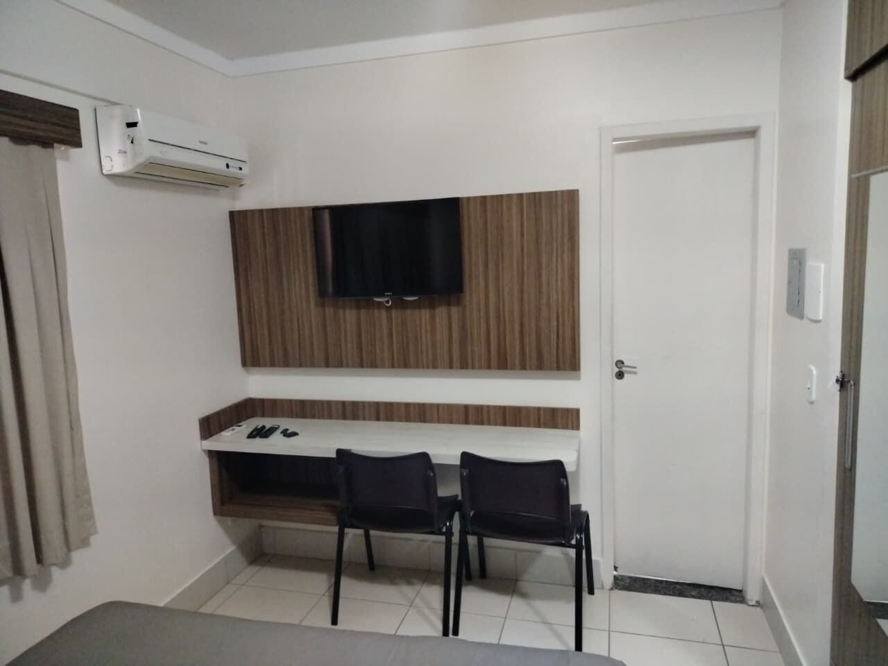 A room with a flat-screen TV mounted on a wooden panel, a white desk with two chairs, and a white door.