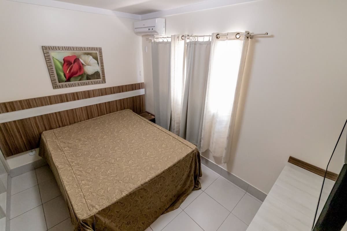 Un dormitorio con cama, aire acondicionado de pared, ventana con cortinas e imagen enmarcada en la pared.