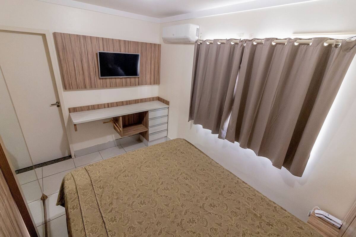 Un dormitorio con cama, televisor montado en un panel de madera, ventana con cortinas y aire acondicionado integrado.
